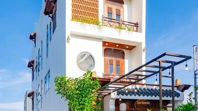 A tall, white, three-story Hoi An building with wooden balconies and lush green plants in front, under a clear blue sky.