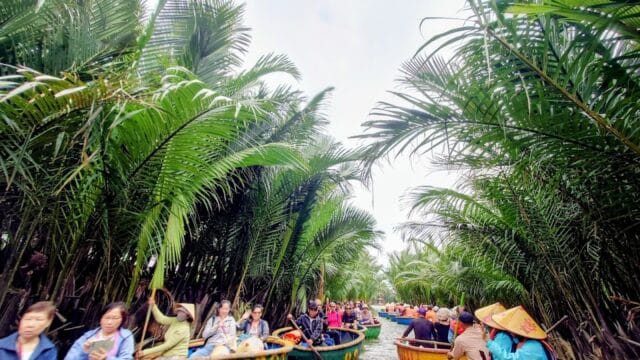 People ride round boats through Hoi An’s lush, green palm forest waterway, wearing hats and colorful clothing under a cloudy sky.