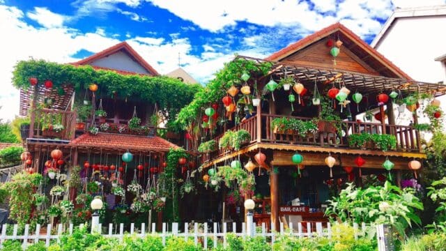 Two wooden houses in Hoi An with lush greenery, colorful lanterns, and a white fence under a blue sky.