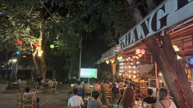 People sit outdoors at night under lanterns in Hoi An, watching a soccer game projected on a screen at a lively café.