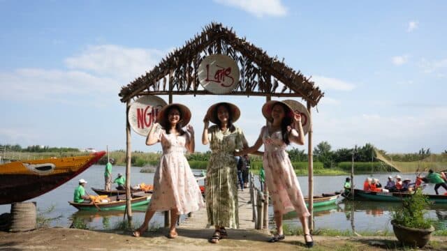 Three women in dresses and hats pose, smiling, under a rustic riverside arch in Hoi An as boats and people bustle behind them.