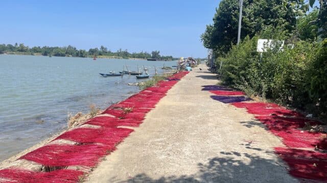 Red incense sticks drying on a riverside path in Hoi An under a clear blue sky, with boats floating nearby.