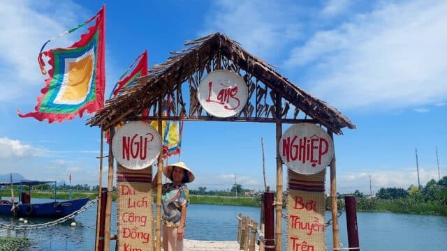 A woman in a conical hat stands under a bamboo gate by a river in Hoi An, with colorful flags and Vietnamese signs.