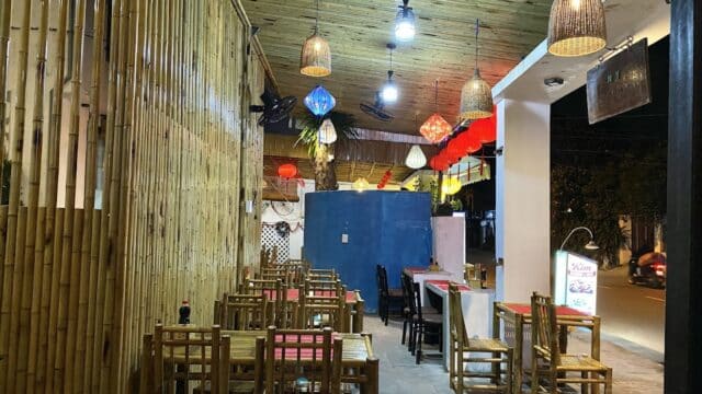 Outdoor Hoi An restaurant with bamboo walls, wooden tables and chairs, colorful lanterns, and warm lighting at night.