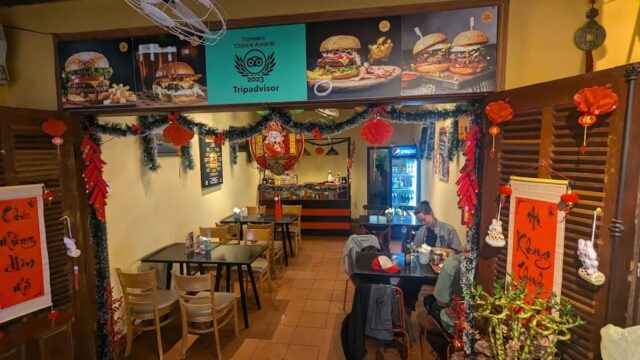 Cozy Hoi An restaurant with festive red decorations, diners enjoying meals, and burger posters above the counter.