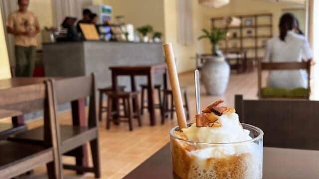 Iced coffee with whipped cream and caramel in a cozy Hoi An café, people chatting near the counter in the background.