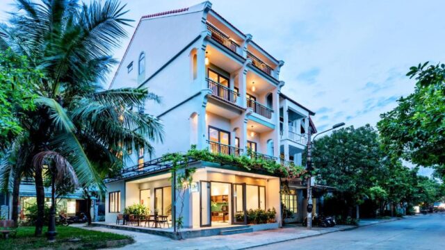Modern three-story building with balconies and large windows, nestled amid greenery in Hoi An on a quiet street with trees and outdoor seating.