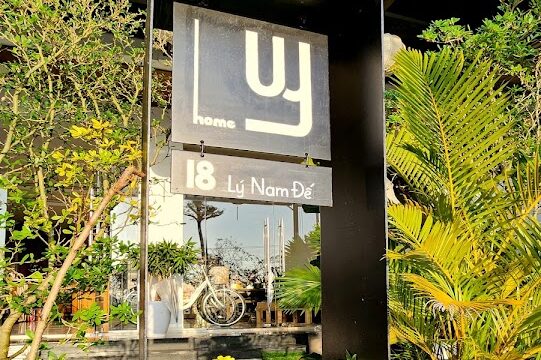 A black sign reading 18 Lý Nam Đế stands amidst yellow flowers and greenery, evoking Hoi An charm before a glass-walled building.