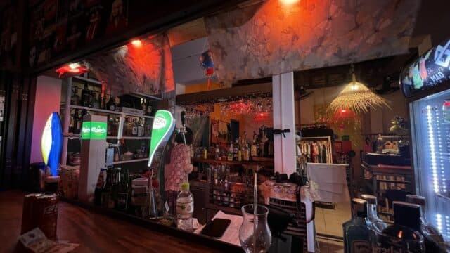 A dimly lit bar in Hoi An with bottles, glasses, red lights, a refrigerator, and clothing racks visible in the background.