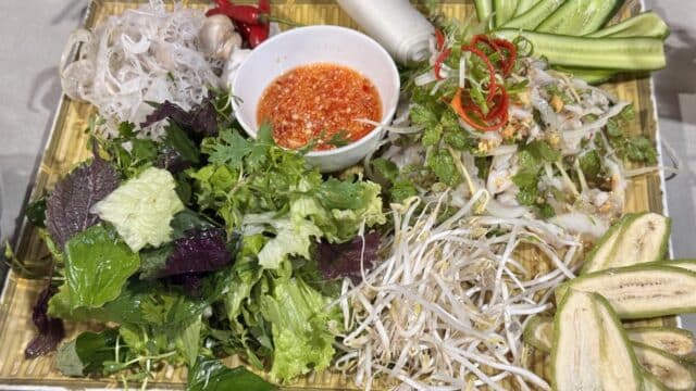 A Hoi An-style platter with greens, cucumber, eggplant, rice noodles, herbs, and dipping sauce in a bowl at the center.