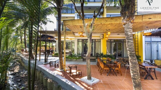 Sunny outdoor restaurant patio with wooden tables and chairs, surrounded by palm trees and a yellow Hoi An building, near a small canal.