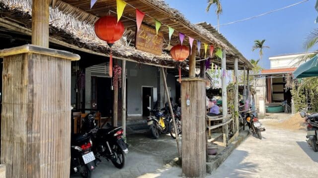 Outdoor cafe in Hoi An with bamboo pillars, colorful flags, red lanterns, and parked motorcycles on a sunny day.