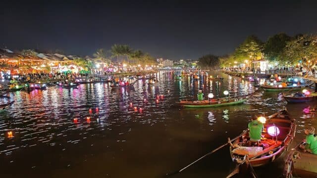 Colorful lantern-lit boats float on Hoi An’s river at night during a lively festival, with crowds along the shore.