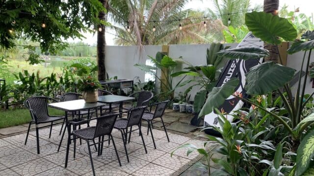 Outdoor patio with a table, six chairs, lush green plants, and a view of Hoi An palm trees and fields in the background.