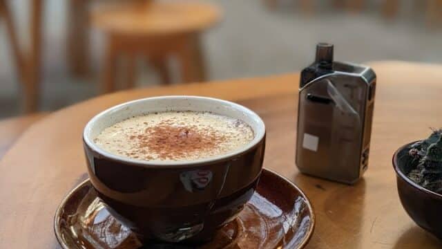 A cappuccino in a brown cup on a saucer sits on a wooden table in Hoi An, with blurred café seating softly in the background.