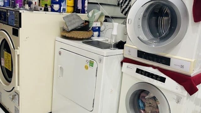 Laundry area in Hoi An with three washing machines, cleaning products, and towels on white tile floor under a staircase.