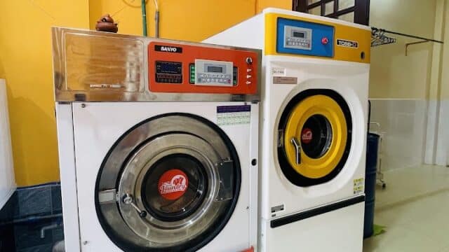 Two industrial washing machines in a brightly lit Hoi An laundry room with yellow walls and detergent on the floor.