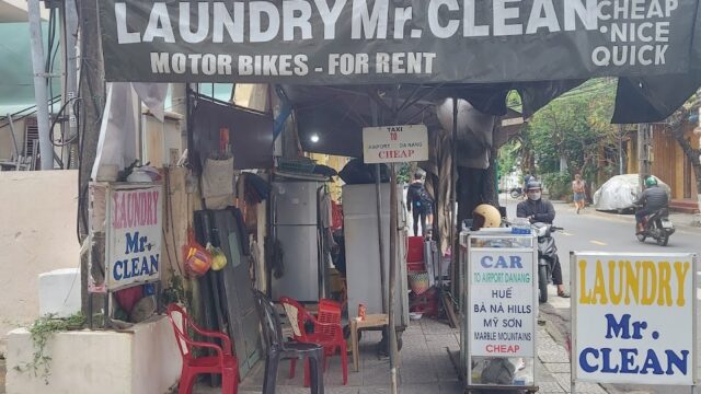 Street view of a laundry shop in Hoi An with signs for laundry, motorbike rental, and car services in Vietnam.