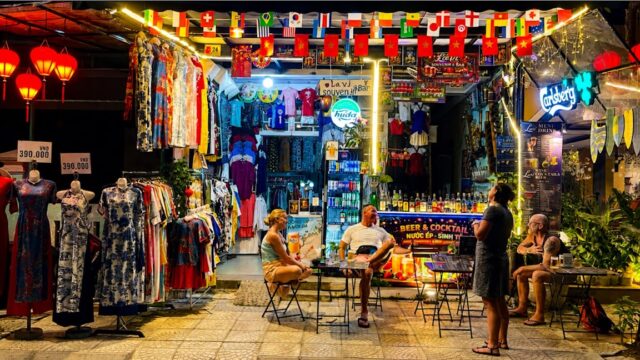 Four people sit at tables outside a brightly lit Hoi An souvenir and clothing shop decorated with international flags.