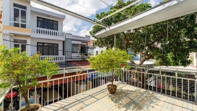 Sunny balcony with potted plants, overlooking a peaceful residential street in Hoi An, framed by charming houses and trees.