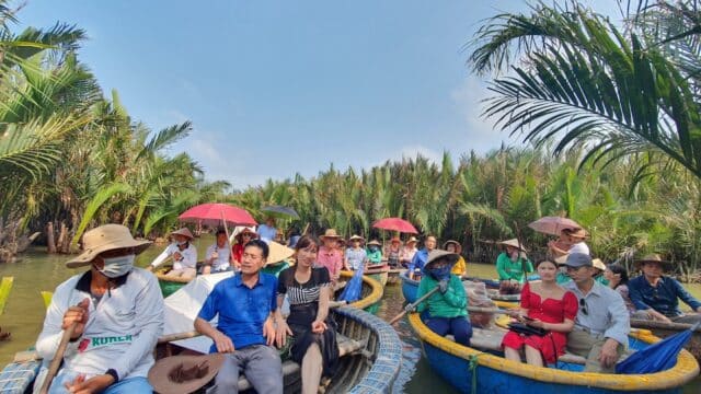 A group of people ride round basket boats through a lush, palm-lined waterway in Hoi An on a sunny day.