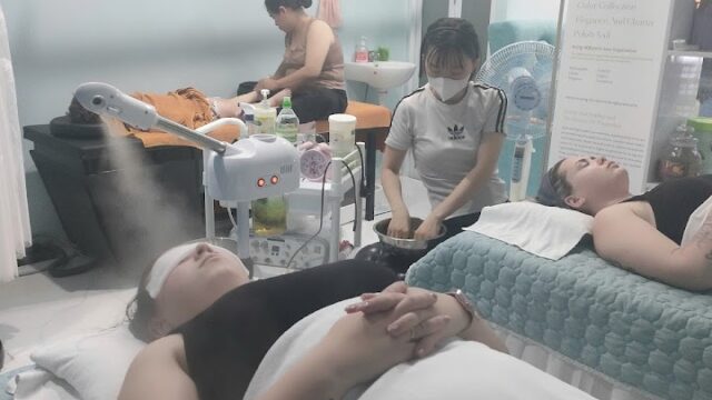 Two people receive facial treatments in a Hoi An spa, as a technician works nearby. Spa equipment and posters adorn the serene room.