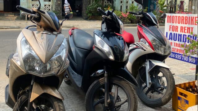 Three parked motorbikes on a sidewalk near a Motorbike for Rent sign in Hoi An on a sunny day.