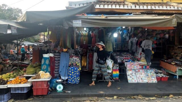 A woman in a hat walks past a colorful Hoi An street market with clothes, fruit, and souvenirs on display.
