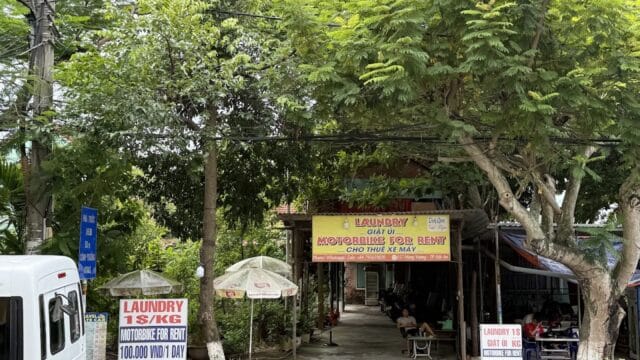 Outdoor laundry and motorbike rental shop in Hoi An, with signs, trees, parked motorbikes, and people sitting at tables along the sidewalk.