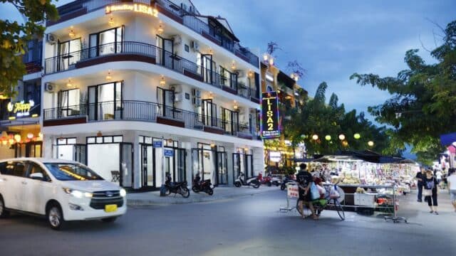A street corner at dusk in Hoi An, with a lit hotel, parked vehicles, and a sidewalk vendor selling items.
