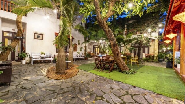 A cozy Hoi An patio with tables, trees, string lights, and a stone path surrounded by greenery and charming buildings.