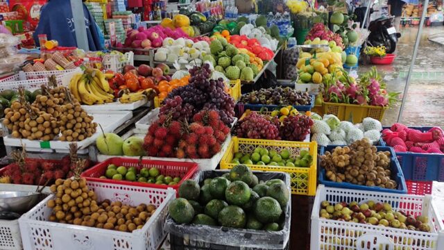 A vibrant Hoi An market stall displays assorted fresh fruits in baskets, including bananas, grapes, avocados, and rambutans.