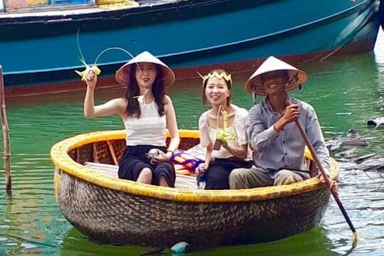 Three people in a round basket boat in Hoi An, smiling with food, wearing conical hats; fishing boats and greenery complete the scene.