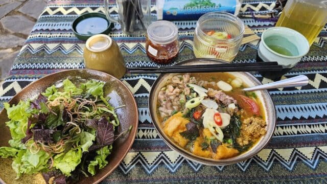 A table set with Hoi An noodle soup, leafy salad, drinks, condiments, and chopsticks rests on a patterned cloth.