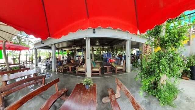 Open-air Hoi An restaurant with wooden benches, tables, and red awnings. Green plants surround the seating area as people dine inside.