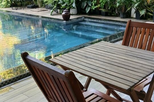 Wooden patio table and chairs beside a clear pool in Hoi An, surrounded by lush tropical plants and trees.