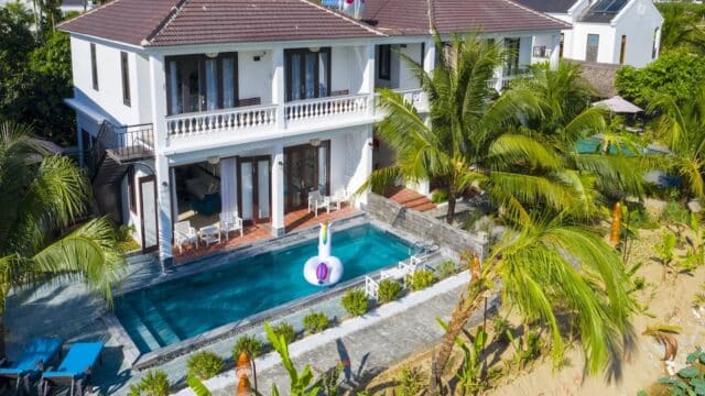 Two-story white villa with a pool, palm trees, and large inflatable swan in Hoi An, just steps from a sandy beach.