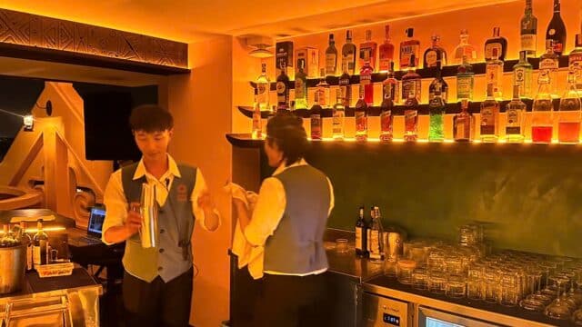 Two bartenders work behind a bar with shelves of illuminated liquor bottles in a warmly lit Hoi An room.