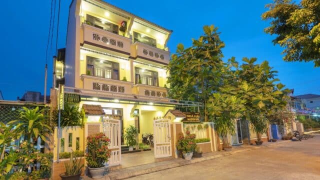 Three-story yellow guesthouse in Hoi An with balconies, potted plants, and warm lighting, nestled among trees at dusk.