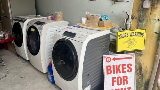 Three washing machines in Hoi An beside signs reading SHOES WASHING and BIKES FOR RENT, with cleaning supplies on top.