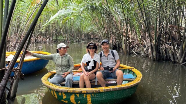 Three people sit in a round boat on a narrow river in Hoi An, surrounded by dense palm trees and lush tropical scenery.