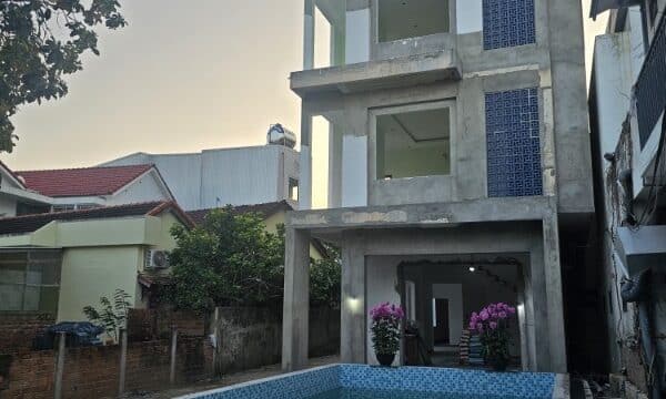 A three-story unfinished building with a tiled pool in front, surrounded by Hoi An houses and plants at sunset.