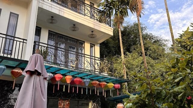 A three-story Hoi An house with balconies, colorful lanterns, tall palms, and a blue sky in the background.