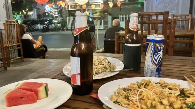 Plates of food, watermelon, and drinks on a table at a Hoi An outdoor restaurant with lanterns and street view.