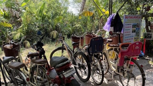 Several bicycles and a red scooter are parked under a blue roof in Hoi An, surrounded by lush green plants and trees.