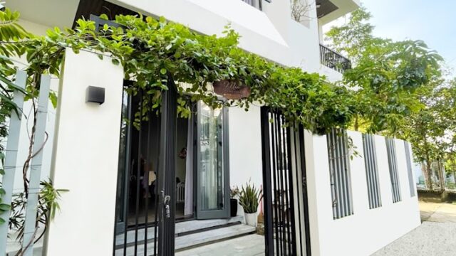 Modern white house with black gate, Hoi An-inspired greenery vines above entrance, and large windows on a sunny day.