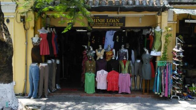 Clothing shop in Hoi An with various mannequins in colorful outfits displayed outside, under a tiled roof storefront.