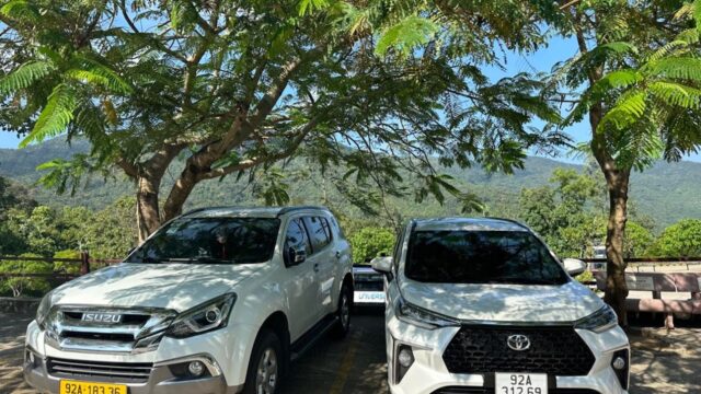 Two white SUVs parked side by side under leafy trees in Hoi An, with mountains and lush greenery visible in the background.