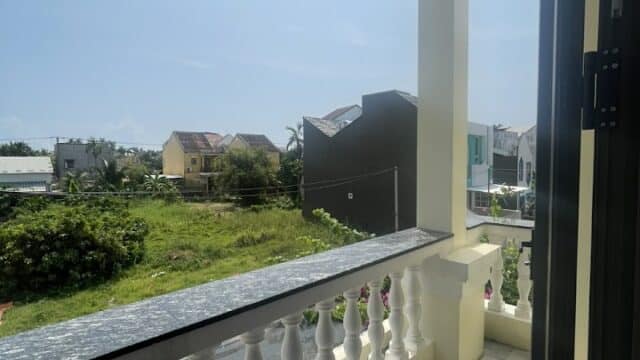 Sunny balcony with white railing overlooks green grass, houses, and a clear blue Hoi An sky in a residential neighborhood.