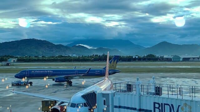 Two airplanes at an airport gate on a rainy day, with mountains and cloudy sky in the background near Hoi An.